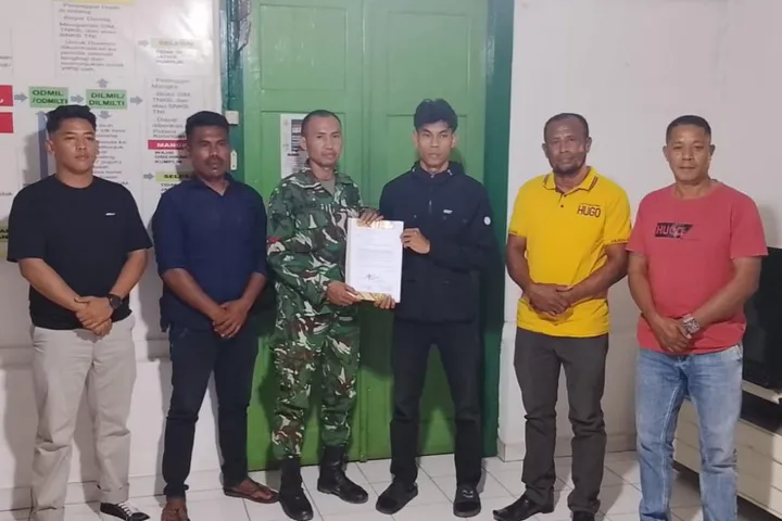 Six men pose indoors as a soldier in camouflage hands a document to a young man in black others watch the handover
